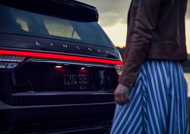 A person is shown near the rear of a 2024 Lincoln Aviator® SUV as the Lincoln Embrace illuminates the rear lights | Irwin Lincoln Laconia in Laconia NH